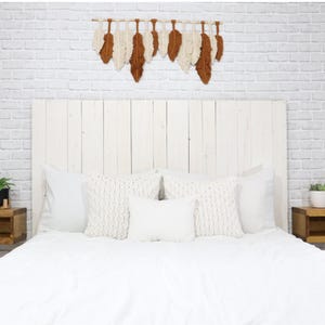 May include: A wall hanging featuring a series of alternating cream and rust-colored fiber feathers suspended from a horizontal wooden dowel. The feathers are arranged in a row, creating a textured, layered appearance against a white brick wall.