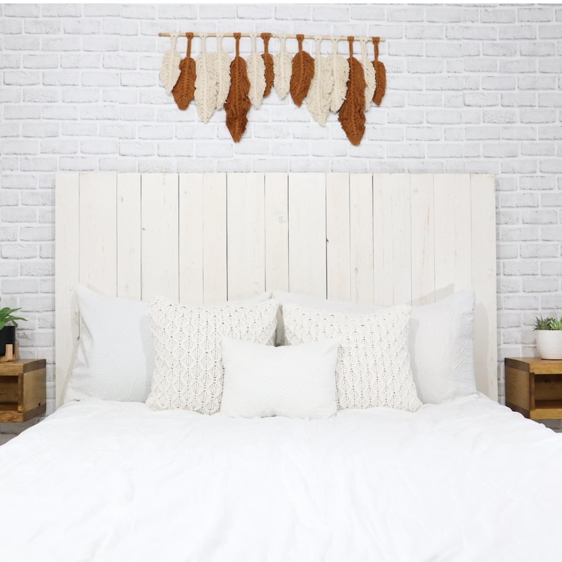 May include: A wall hanging featuring a series of alternating cream and rust-colored fiber feathers suspended from a horizontal wooden dowel. The feathers are arranged in a row, creating a textured, layered appearance against a white brick wall.