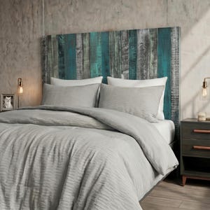 May include: A headboard made of vertical wooden planks in shades of teal, grey, and dark brown. The bed is dressed in a light grey comforter with a textured, rippled surface and two matching pillows. The headboard features a weathered finish.
