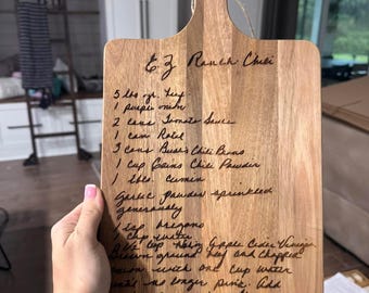 May include: A wooden cutting board with a handwritten recipe for "6.3 Ranch Chili" on it. The recipe includes ingredients like 5 lbs of ground beef, 1 purple onion, 2 cans of tomato sauce, 1 can of Rotel, 3 cans of Bush's Chili Beans, 1 cup of beans chili powder, 1 tbs of cumin, garlic powder sprinkled generously, 1 cup oregano, 1/2 cup white apple cider vinegar, 1/2 cup chopped green onion, 1 cup water, and mix until no longer pink. Add beans and tomato sauce, Rotel and beans and 1/2 cup vinegar.
