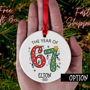 May include: A white ceramic Christmas ornament with the text "THE YEAR OF 67" in red and green, decorated with string lights and stars. The name "ELTON -2025" is printed below. A red ribbon is attached for hanging.