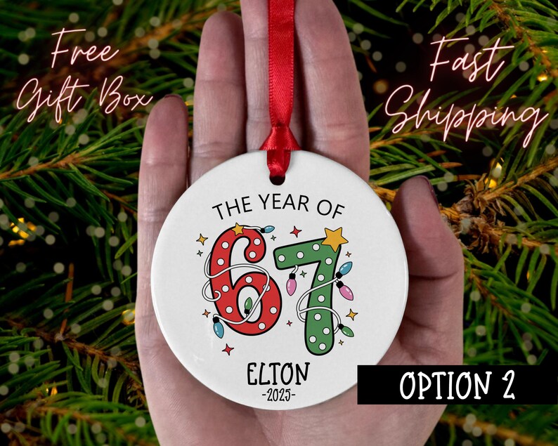 May include: A white ceramic Christmas ornament with the text "THE YEAR OF 67" in red and green, decorated with string lights and stars. The name "ELTON -2025" is printed below. A red ribbon is attached for hanging.