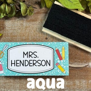 May include: An aqua-colored teacher nameplate with the name "MRS. HENDERSON" in a white frame, decorated with school supplies. A black chalkboard eraser sits above the nameplate. The word "aqua" is at the bottom.