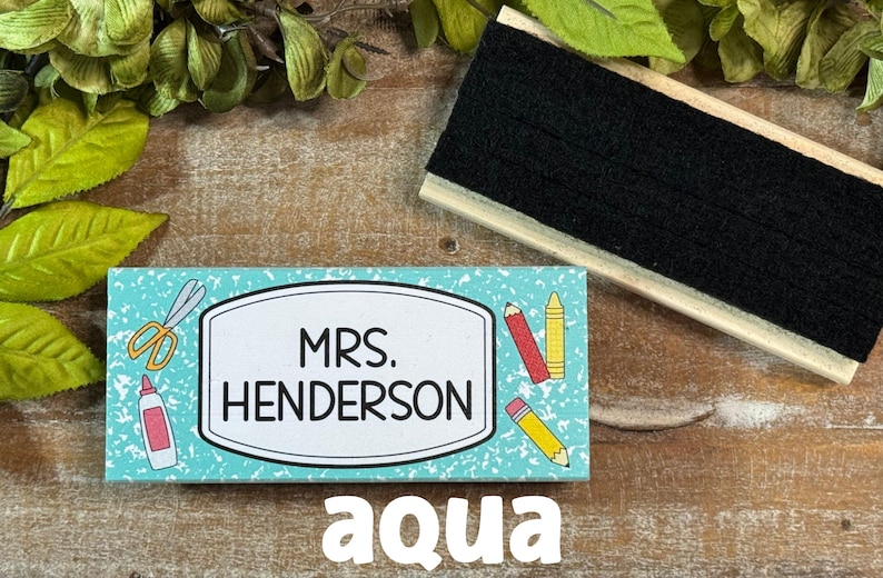 May include: An aqua-colored teacher nameplate with the name "MRS. HENDERSON" in a white frame, decorated with school supplies. A black chalkboard eraser sits above the nameplate. The word "aqua" is at the bottom.