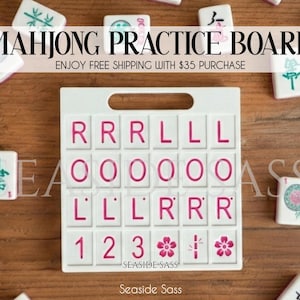 May include: White Mahjong practice board with pink lettering and a handle. The board has spaces for tiles and the text "MAHJONG PRACTICE BOARD". The board is surrounded by Mahjong tiles on a wooden surface.