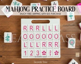 May include: White Mahjong practice board with pink lettering and a handle. The board has spaces for tiles and the text "MAHJONG PRACTICE BOARD". The board is surrounded by Mahjong tiles on a wooden surface.