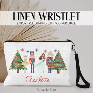 May include: White linen wristlet with a black strap. The wristlet features a watercolor illustration of a Nutcracker, a ballerina, and two Christmas trees. The text "Charlotte" is printed on the front of the wristlet.