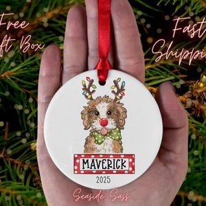 May include: A white ceramic Christmas ornament featuring a dog with reindeer antlers and a red nose. The dog has a green bow tie and the name "MAVERICK" is displayed on a red and white banner. The year "2025" is also shown.