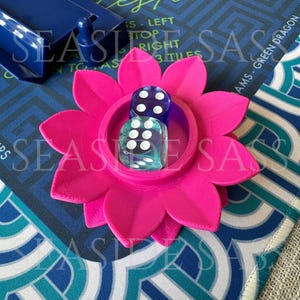 May include: A bright pink, flower-shaped dice holder with two dice inside. The dice are clear with white dots. The holder sits on a patterned game board with text that reads "Leston" and "AMS - GREEN DRAGON".