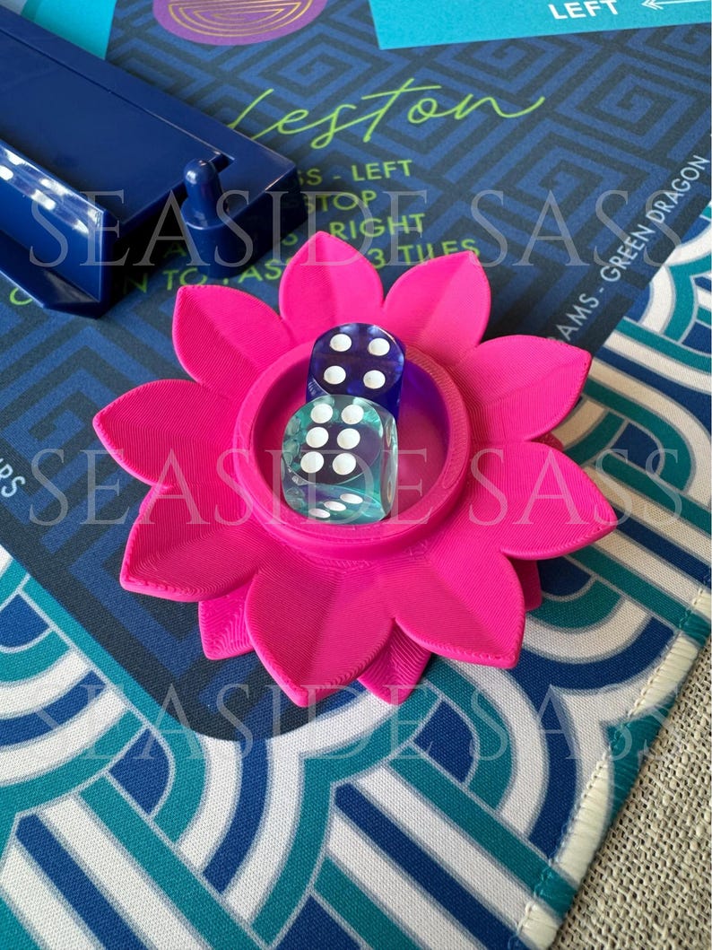 May include: A bright pink, flower-shaped dice holder with two dice inside. The dice are clear with white dots. The holder sits on a patterned game board with text that reads "Leston" and "AMS - GREEN DRAGON".