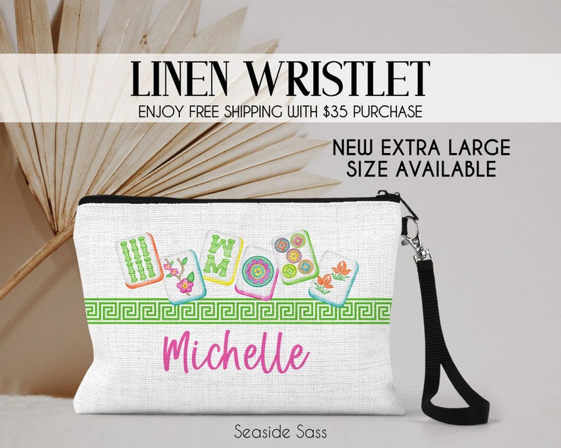 May include: A white linen wristlet with a black zipper and a black strap. The wristlet has a colorful design of mahjong tiles with the name "Michelle" embroidered on the front. The text "LINEN WRISTLET" and "ENJOY FREE SHIPPING WITH $35 PURCHASE" is at the top of the image. The text "NEW EXTRA LARGE SIZE AVAILABLE" is in the middle of the image. The text "Seaside Sass" is at the bottom of the image.
