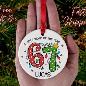May include: A white ceramic Christmas ornament with a red ribbon. The ornament features the numbers 6 and 7 in red and green, with the text "2025 WORD OF THE YEAR" and the name "LUCAS". The numbers are decorated with Christmas lights.