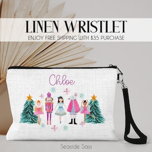 May include: White linen wristlet with a black zipper and wrist strap. The wristlet features a watercolor design of Christmas characters, including a ballerina, nutcracker, and mouse. The name "Chloe" is written in cursive. The text "Linen Wristlet" is at the top.