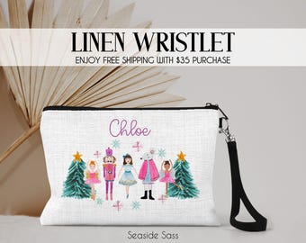 May include: White linen wristlet with a black zipper and wrist strap. The wristlet features a watercolor design of Christmas characters, including a ballerina, nutcracker, and mouse. The name "Chloe" is written in cursive. The text "Linen Wristlet" is at the top.
