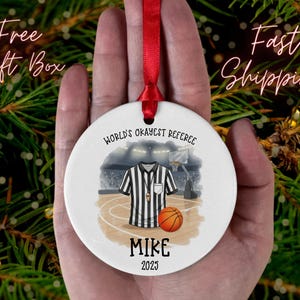 May include: A white ceramic ornament with a red ribbon, featuring a basketball referee design. The ornament has the text "World's Okayest Referee" and "Mike 2025". The design includes a referee shirt, basketball, and court.