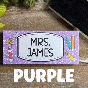 May include: A rectangular purple name tag with the text "MRS. JAMES" in a white oval. The tag is decorated with school supplies like scissors, glue, and pencils. The word "PURPLE" is written in large white letters below.