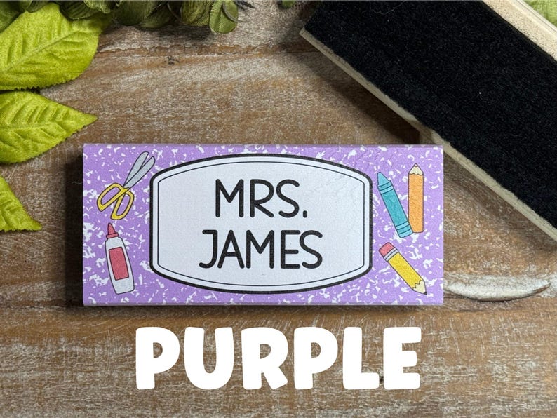 May include: A rectangular purple name tag with the text "MRS. JAMES" in a white oval. The tag is decorated with school supplies like scissors, glue, and pencils. The word "PURPLE" is written in large white letters below.
