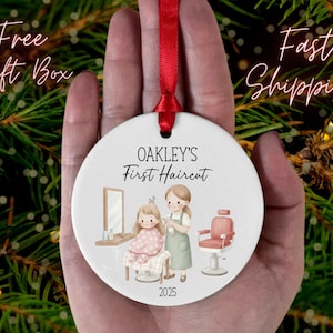 May include: A white ceramic ornament with a red ribbon, featuring a salon scene with the text "OAKLEY'S First Haircut" and the year 2025. The ornament depicts a child getting a haircut, with a stylist and salon chair.