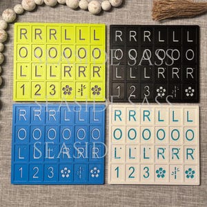 Może przedstawiać: Cztery kolorowe uchwyty na kafelki Scrabble w kolorze ż&oacute;łtym, czarnym, niebieskim i białym. Każdy uchwyt ma kafelki z literami, cyframi i kwiatowymi wzorami. Uchwyty są ułożone na neutralnej powierzchni.