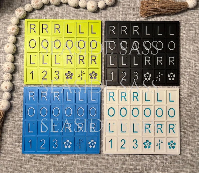 Może przedstawiać: Cztery kolorowe uchwyty na kafelki Scrabble w kolorze ż&oacute;łtym, czarnym, niebieskim i białym. Każdy uchwyt ma kafelki z literami, cyframi i kwiatowymi wzorami. Uchwyty są ułożone na neutralnej powierzchni.