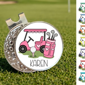 May include: A silver golf divot tool with a circular design featuring a pink golf cart, golf bag, golf ball, and the name "KAREN" in black text. The golf cart has heart details. The tool is set on a green grass background.