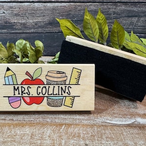 May include: A wooden desk nameplate with the name "MRS. COLLINS" and school-themed illustrations, including a pencil, apple, coffee cup, and ruler. A black chalkboard eraser rests beside the nameplate.