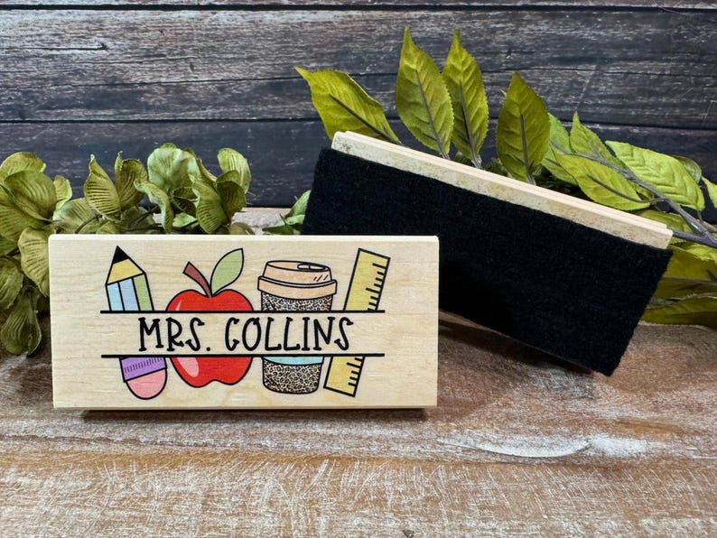 May include: A wooden desk nameplate with the name "MRS. COLLINS" and school-themed illustrations, including a pencil, apple, coffee cup, and ruler. A black chalkboard eraser rests beside the nameplate.