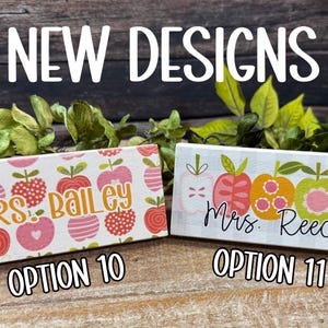 May include: Two rectangular wooden signs with apple designs. One sign says "Mrs. Bailey" and the other says "Mrs. Reece". The signs are decorated with pink, red, and orange apples. The text "NEW DESIGNS" is at the top.
