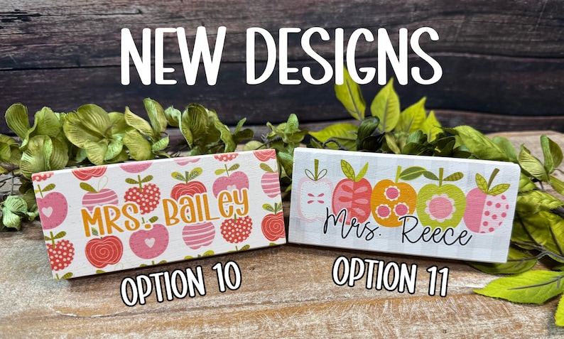 May include: Two rectangular wooden signs with apple designs. One sign says "Mrs. Bailey" and the other says "Mrs. Reece". The signs are decorated with pink, red, and orange apples. The text "NEW DESIGNS" is at the top.