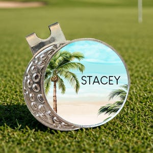 May include: A silver golf ball marker with a tropical beach scene and the name "STACEY" in white lettering. The marker is on a green golf course.