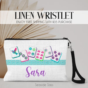 K&ouml;nnte beinhalten: Eine wei&szlig;e Leinen-Handgelenktasche mit einem Grafikdruck von sechs Mahjong-Steinen, darunter einer mit einem Kolibri, und einer t&uuml;rkisfarbenen griechischen Bord&uuml;re. Der Name Sara ist in violetter Schrift darunter gedruckt. Ein schwarzes Band ist seitlich befestigt.