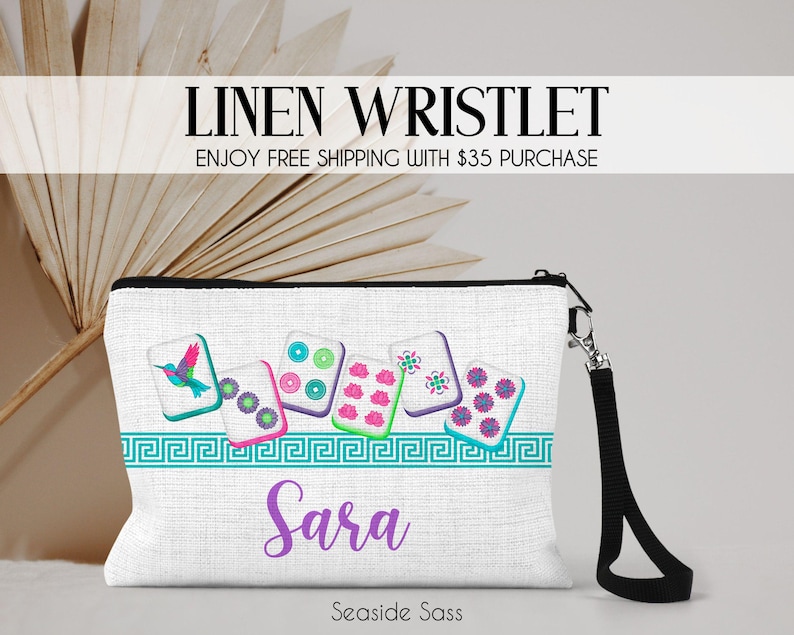 K&ouml;nnte beinhalten: Eine wei&szlig;e Leinen-Handgelenktasche mit einem Grafikdruck von sechs Mahjong-Steinen, darunter einer mit einem Kolibri, und einer t&uuml;rkisfarbenen griechischen Bord&uuml;re. Der Name Sara ist in violetter Schrift darunter gedruckt. Ein schwarzes Band ist seitlich befestigt.