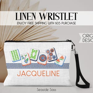 K&ouml;nnte beinhalten: Eine wei&szlig;e Leinen-Handgelenktasche mit schwarzer Handschlaufe und einem Grafikdruck von sechs bunten Mahjong-Steinen &uuml;ber einer blauen M&auml;anderbord&uuml;re. Der Name JACQUELINE ist in orangefarbenen Gro&szlig;buchstaben unter der Bord&uuml;re aufgedruckt.