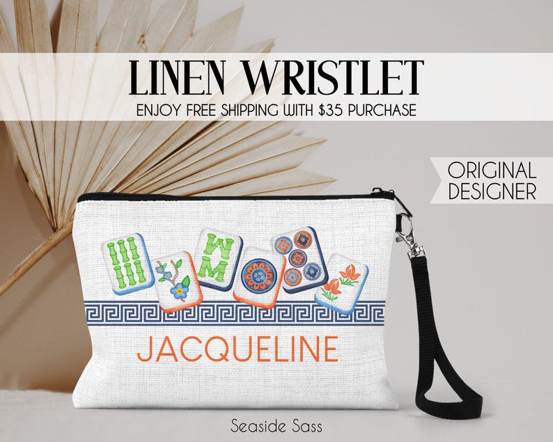 K&ouml;nnte beinhalten: Eine wei&szlig;e Leinen-Handgelenktasche mit schwarzer Handschlaufe und einem Grafikdruck von sechs bunten Mahjong-Steinen &uuml;ber einer blauen M&auml;anderbord&uuml;re. Der Name JACQUELINE ist in orangefarbenen Gro&szlig;buchstaben unter der Bord&uuml;re aufgedruckt.