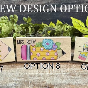 May include: Three wooden teacher desk nameplates with pencil and apple designs. Each nameplate features a different design and name, with options 7, 8, and 9 labeled below. The nameplates are painted with bright colors and are displayed on a wooden surface.