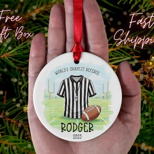 May include: A white ceramic ornament with a red ribbon, featuring a referee's uniform and a football. Text reads "World's Okayest Referee," the name "Rodger," and the year "2025."