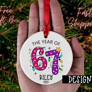 May include: A white ceramic ornament with a red ribbon, featuring the text "THE YEAR OF 67 RILEY -2023". The numbers are decorated with pink polka dots and purple polka dots, with colorful lights and stars. The design is labeled "DESIGN 5".