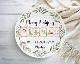 May include: White ceramic plate with a festive design. The plate features the text "Merry Mahjong" and illustrations of Christmas-themed Mahjong tiles. A green and red floral border encircles the plate, with the name "Marilyn" at the bottom.