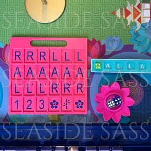 May include: A pink Scrabble tile holder with blue letter tiles, a blue tile holder with the word "ALLAR", a pink flower with a die, and a background with floral and geometric patterns. The text "SEASIDE SASS" is visible.