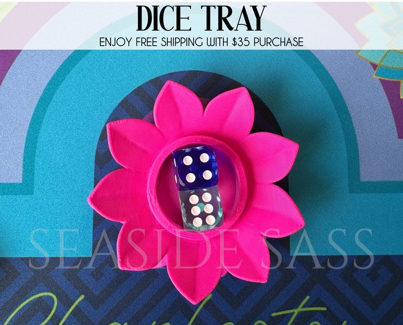 May include: A hot pink flower-shaped dice tray with two dice inside. The dice are blue and clear with white dots. The text "DICE TRAY" is at the top of the image, and "SEASIDE SASS" is at the bottom.