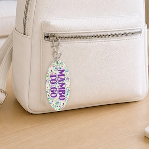 May include: An oval-shaped keychain with the text "MAMBO TO GO" in white and purple. The keychain is attached to a white backpack with a silver zipper. The keychain has a pattern of colorful shapes and dots.