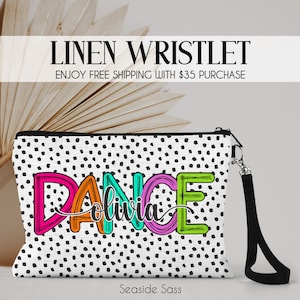 May include: A white linen wristlet with a black polka dot pattern and the word "DANCE" in colorful letters with the name "Olivia" written in cursive underneath. The wristlet has a black strap.