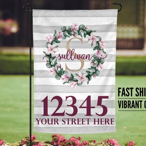 May include: A personalized garden flag with a gray and white striped background. A wreath of pink flowers and green leaves surrounds a gold letter "S" with the name "sullivan" written in cursive. The number "12345" and the text "YOUR STREET HERE" are printed in burgundy.