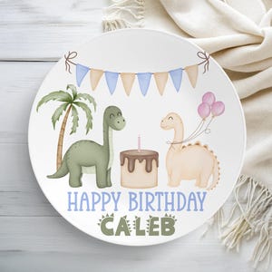 Könnte beinhalten: Ein weißer Keramikteller mit Geburtstagsthema. Der Teller hat eine Cartoon-Illustration von zwei Dinosauriern, einem Kuchen mit einer Kerze und Luftballons. Der Text "HAPPY BIRTHDAY CALEB" ist in Blau und Grün auf dem Teller geschrieben.