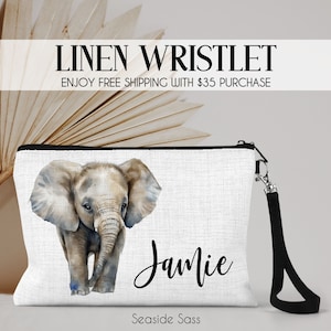 May include: A white linen wristlet with a black zipper and a black wrist strap. The wristlet features a watercolor illustration of a baby elephant and the name "Jamie" printed in black cursive. The text "Seaside Sass" is printed in black at the bottom of the wristlet.
