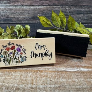 May include: A wooden desk nameplate with the name "Mrs. Murphy" in black script. The nameplate is decorated with a floral design and the words "Teach them, love them, watch them grow." A black eraser is next to the nameplate.