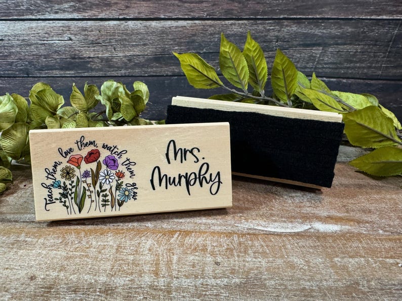 May include: A wooden desk nameplate with the name "Mrs. Murphy" in black script. The nameplate is decorated with a floral design and the words "Teach them, love them, watch them grow." A black eraser is next to the nameplate.