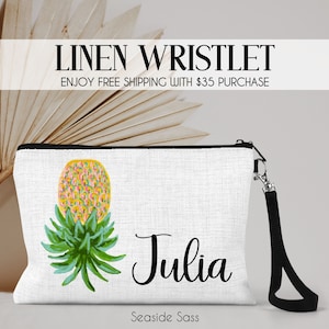 May include: A white linen wristlet with a black zipper and a black strap. The wristlet features a colorful pineapple design and the name "Julia" in black cursive writing. The text "Seaside Sass" is printed in black at the bottom of the wristlet.