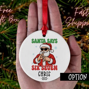 May include: A white ceramic Christmas ornament with a red ribbon. The ornament features a cartoon Santa Claus wearing sunglasses and the text "SANTA SAYS SIX SEVEN CHRIS -2025". The ornament is held against a background of green pine needles.