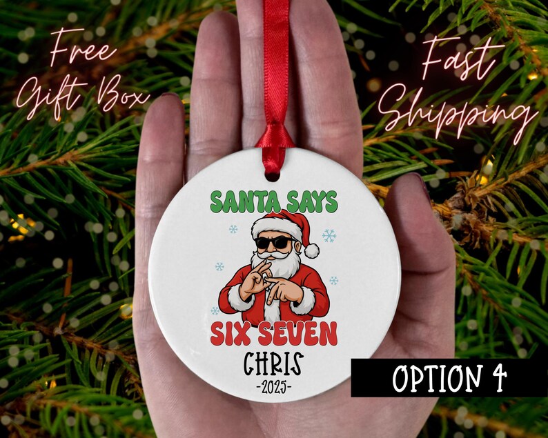 May include: A white ceramic Christmas ornament with a red ribbon. The ornament features a cartoon Santa Claus wearing sunglasses and the text "SANTA SAYS SIX SEVEN CHRIS -2025". The ornament is held against a background of green pine needles.